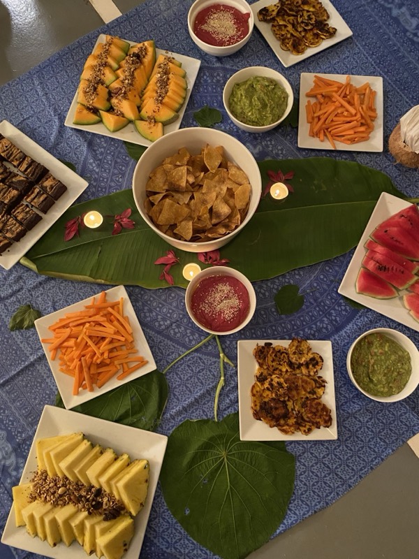 Mesa servida vista desde arriba: fuentes de verduras, frutas y salsas sobre hojas grandes.